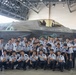 A large group of what appears to be Korean military cadets or students in light blue/grey uniforms pose for a group photo in front of an F-35B fighter jet inside a hangar. The aircraft bears the markings 'VMFA-121', identifying it as belonging to Marine Fighter Attack Squadron 121, a U.S. Marine Corps unit. Two U.S. military personnel — one in camouflage utilities and one in a flight suit — are also present in the photo.