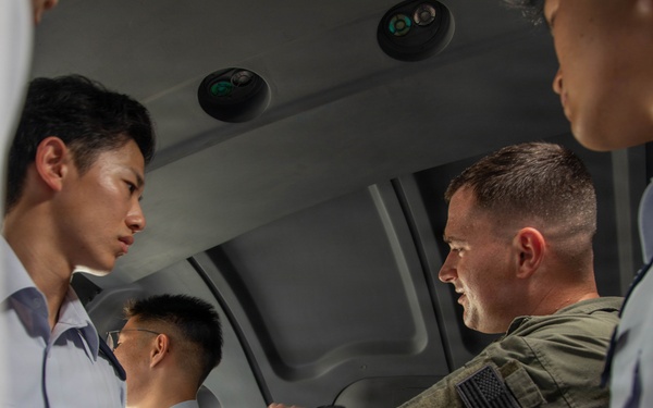 A U.S. military personnel member in olive green flight suit with an American flag patch is briefing or instructing a group of Asian military personnel wearing light blue uniforms with wing insignia. The setting appears to be inside a military aircraft or simulator cockpit/cabin. The interaction suggests a joint training or exchange program between U.S. and allied air force personnel.