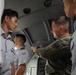 A U.S. military personnel member in olive green flight suit with an American flag patch is briefing or instructing a group of Asian military personnel wearing light blue uniforms with wing insignia. The setting appears to be inside a military aircraft or simulator cockpit/cabin. The interaction suggests a joint training or exchange program between U.S. and allied air force personnel.