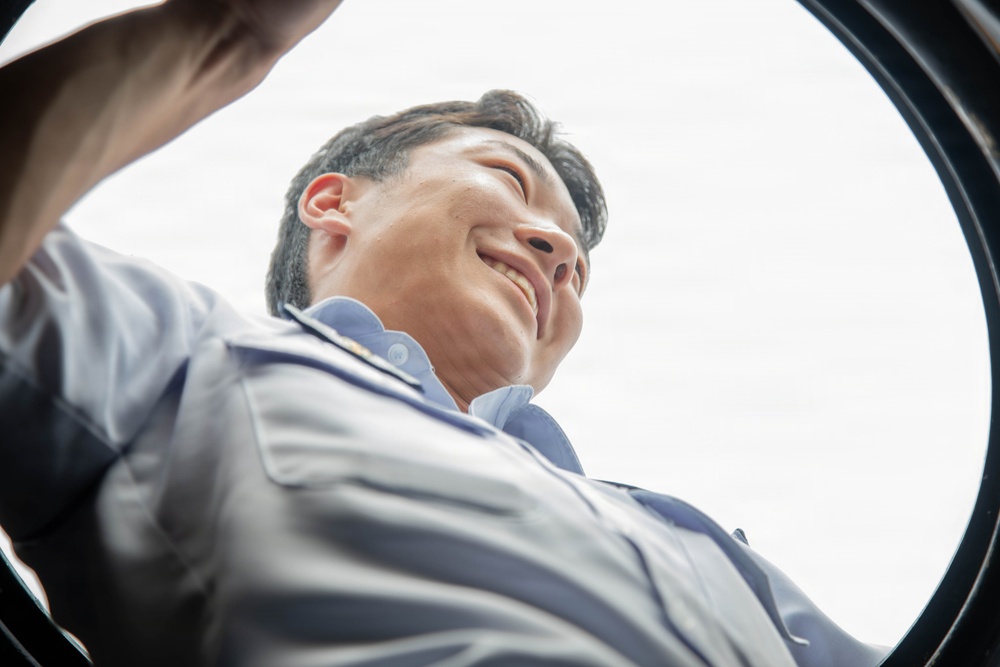 The image is taken from a low-angle perspective looking upward through a circular opening, likely the drum of a washing machine or similar cylindrical appliance. A smiling man wearing a light blue work uniform is peering down into the opening. The unusual vantage point creates a dramatic, fish-eye-like composition with the sky visible in the background.