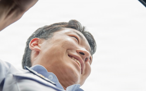 The image is taken from a low-angle perspective looking upward through a circular opening, likely the drum of a washing machine or similar cylindrical appliance. A smiling man wearing a light blue work uniform is peering down into the opening. The unusual vantage point creates a dramatic, fish-eye-like composition with the sky visible in the background.