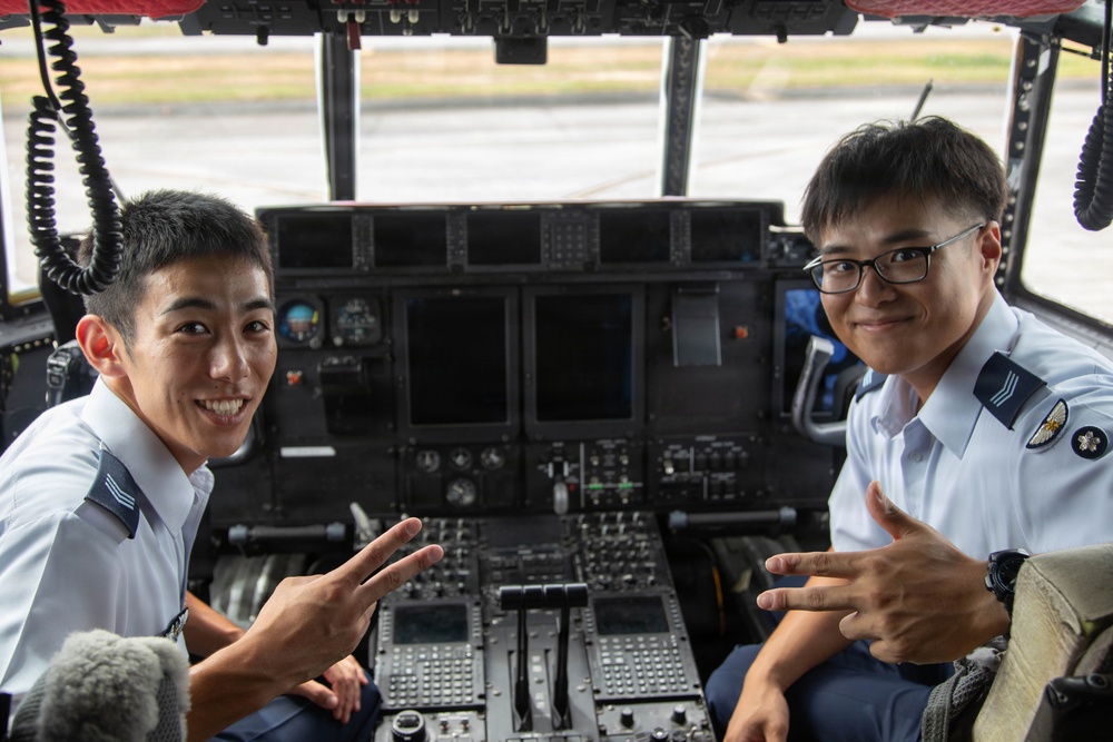 Two uniformed military aviation personnel are seated in the cockpit of a large aircraft, posing for a photo with peace/victory hand signs. Both wear light blue uniforms with rank insignia visible on their shoulders. The cockpit features an extensive instrument panel with multiple displays, switches, and controls consistent with a large military transport aircraft.
