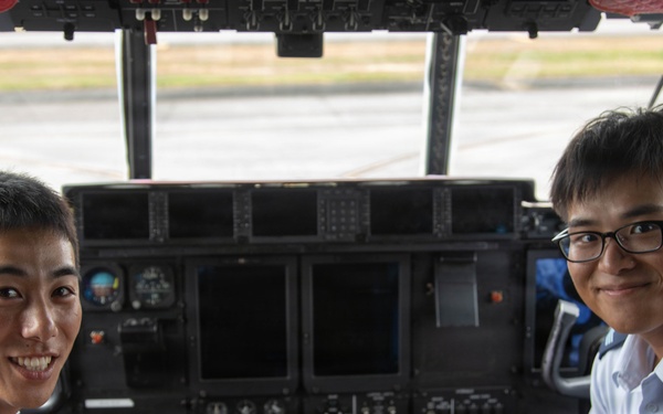 Two uniformed military aviation personnel are seated in the cockpit of a large aircraft, posing for a photo with peace/victory hand signs. Both wear light blue uniforms with rank insignia visible on their shoulders. The cockpit features an extensive instrument panel with multiple displays, switches, and controls consistent with a large military transport aircraft.