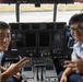 Two uniformed military aviation personnel are seated in the cockpit of a large aircraft, posing for a photo with peace/victory hand signs. Both wear light blue uniforms with rank insignia visible on their shoulders. The cockpit features an extensive instrument panel with multiple displays, switches, and controls consistent with a large military transport aircraft.