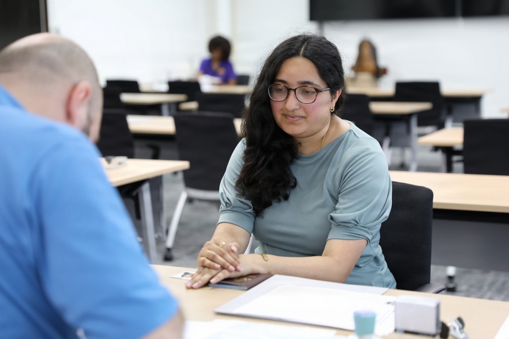 A young woman wearing glasses and a teal top is seated at a table in what appears to be a modern classroom or conference room, engaged in a one-on-one conversation or meeting with a person in a blue shirt whose back is to the camera. She has documents and what appears to be a passport or booklet on the table in front of her. In the background, at least one other person is visible seated at a separate table.