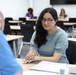 A young woman wearing glasses and a teal top is seated at a table in what appears to be a modern classroom or conference room, engaged in a one-on-one conversation or meeting with a person in a blue shirt whose back is to the camera. She has documents and what appears to be a passport or booklet on the table in front of her. In the background, at least one other person is visible seated at a separate table.