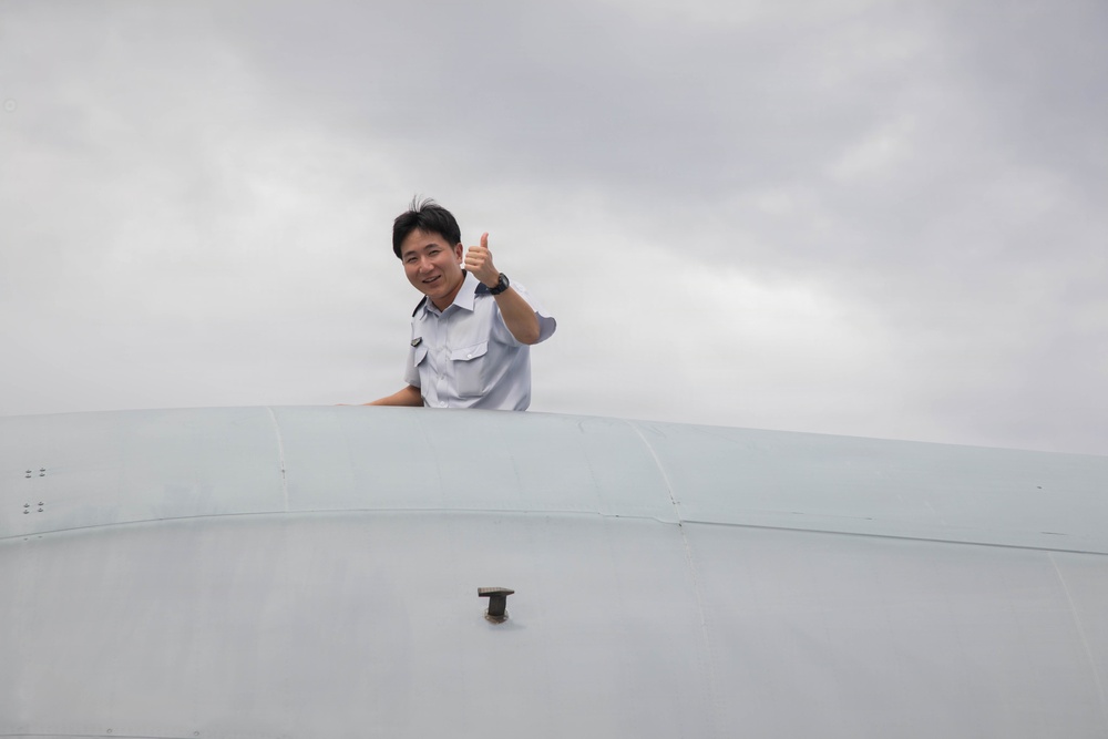 A man in a light blue uniform shirt is visible from the shoulders up, appearing to emerge from or lean over a large white curved surface, likely an aircraft fuselage or similar aviation structure. He is smiling and giving a thumbs-up gesture toward the camera. The background shows an overcast, cloudy sky.