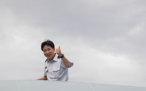 A man in a light blue uniform shirt is visible from the shoulders up, appearing to emerge from or lean over a large white curved surface, likely an aircraft fuselage or similar aviation structure. He is smiling and giving a thumbs-up gesture toward the camera. The background shows an overcast, cloudy sky.