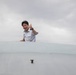 A man in a light blue uniform shirt is visible from the shoulders up, appearing to emerge from or lean over a large white curved surface, likely an aircraft fuselage or similar aviation structure. He is smiling and giving a thumbs-up gesture toward the camera. The background shows an overcast, cloudy sky.