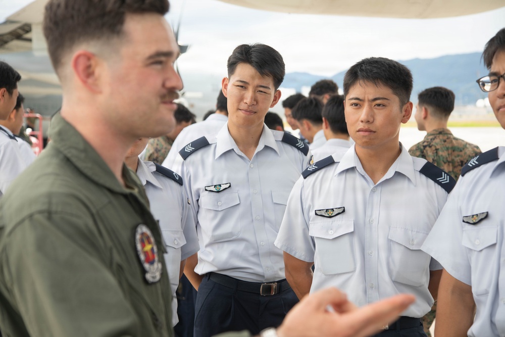 A military briefing or exchange is taking place on an airfield tarmac, where a Western military aviator in an olive green flight suit is speaking to a group of Asian military personnel in white dress uniforms. The setting appears to be a joint military exercise or base visit, with additional personnel visible in the background including individuals in camouflage uniforms. The interaction suggests an international military cooperation or exchange program.