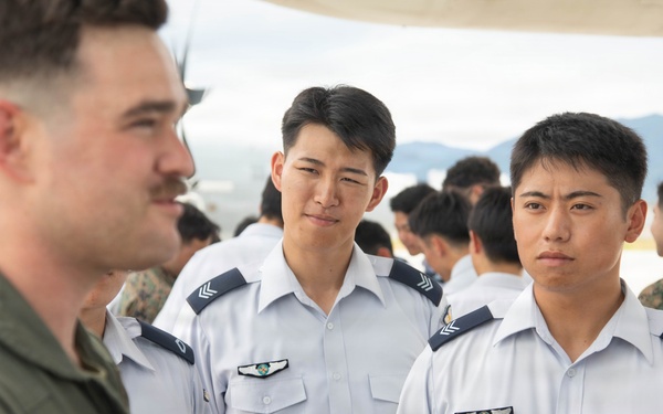 A military briefing or exchange is taking place on an airfield tarmac, where a Western military aviator in an olive green flight suit is speaking to a group of Asian military personnel in white dress uniforms. The setting appears to be a joint military exercise or base visit, with additional personnel visible in the background including individuals in camouflage uniforms. The interaction suggests an international military cooperation or exchange program.