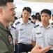 A military briefing or exchange is taking place on an airfield tarmac, where a Western military aviator in an olive green flight suit is speaking to a group of Asian military personnel in white dress uniforms. The setting appears to be a joint military exercise or base visit, with additional personnel visible in the background including individuals in camouflage uniforms. The interaction suggests an international military cooperation or exchange program.