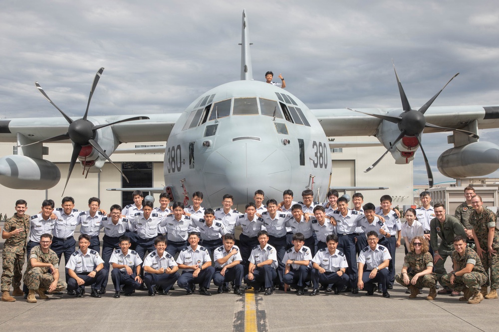 A large group of military personnel pose for a formal group photograph in front of a turboprop military transport aircraft bearing the number '380' on its nose. The group consists primarily of individuals in light blue/grey dress uniforms, flanked by a smaller number of personnel in camouflage combat uniforms. One individual is visible standing atop the aircraft's fuselage.
