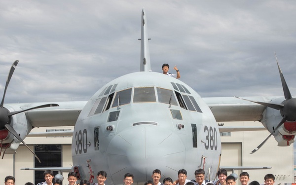A large group of military personnel pose for a formal group photograph in front of a turboprop military transport aircraft bearing the number '380' on its nose. The group consists primarily of individuals in light blue/grey dress uniforms, flanked by a smaller number of personnel in camouflage combat uniforms. One individual is visible standing atop the aircraft's fuselage.