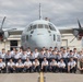 A large group of military personnel pose for a formal group photograph in front of a turboprop military transport aircraft bearing the number '380' on its nose. The group consists primarily of individuals in light blue/grey dress uniforms, flanked by a smaller number of personnel in camouflage combat uniforms. One individual is visible standing atop the aircraft's fuselage.