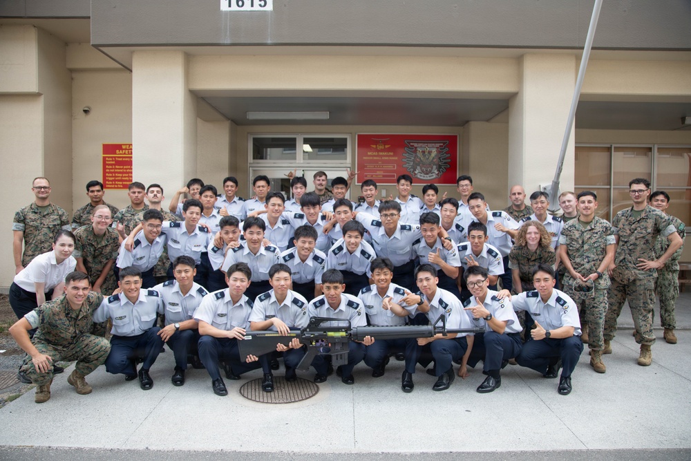 A large group photo taken in front of building number 1615, showing a mix of military personnel in camouflage uniforms and individuals in white dress uniforms with dark trousers, likely cadets or students from an Asian military academy. The group is posing together with a rifle displayed prominently in the front center, suggesting a joint military exchange or training event. A weapon safety sign is visible on the left side of the building entrance.