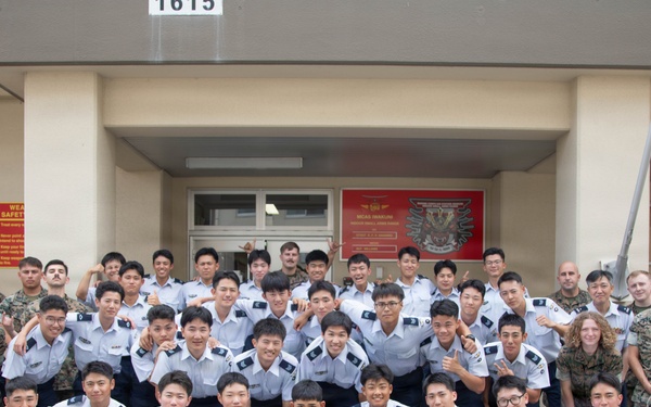 A large group photo taken in front of building number 1615, showing a mix of military personnel in camouflage uniforms and individuals in white dress uniforms with dark trousers, likely cadets or students from an Asian military academy. The group is posing together with a rifle displayed prominently in the front center, suggesting a joint military exchange or training event. A weapon safety sign is visible on the left side of the building entrance.