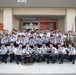 A large group photo taken in front of building number 1615, showing a mix of military personnel in camouflage uniforms and individuals in white dress uniforms with dark trousers, likely cadets or students from an Asian military academy. The group is posing together with a rifle displayed prominently in the front center, suggesting a joint military exchange or training event. A weapon safety sign is visible on the left side of the building entrance.