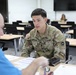 A U.S. Army soldier named 'English' is seated at a table in what appears to be a classroom or conference room, engaged in a discussion or meeting with a civilian in a blue shirt. The soldier is wearing the Operational Camouflage Pattern (OCP) uniform with a U.S. flag patch and U.S. Army tape visible. Documents and papers are spread on the table between them, suggesting an administrative, counseling, or briefing session.