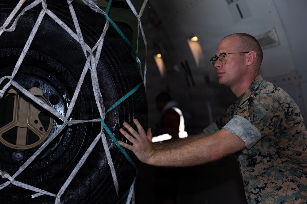 A U.S. Marine in digital camouflage (MARPAT) uniform is inspecting or securing a large tire/wheel assembly that is restrained with a white cargo net inside what appears to be a military aircraft cargo hold. The Marine is wearing glasses and is physically touching the tire, likely conducting a load inspection or securing check. The setting suggests a military airlift or cargo loading operation.