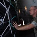 A U.S. Marine in digital camouflage (MARPAT) uniform is inspecting or securing a large tire/wheel assembly that is restrained with a white cargo net inside what appears to be a military aircraft cargo hold. The Marine is wearing glasses and is physically touching the tire, likely conducting a load inspection or securing check. The setting suggests a military airlift or cargo loading operation.