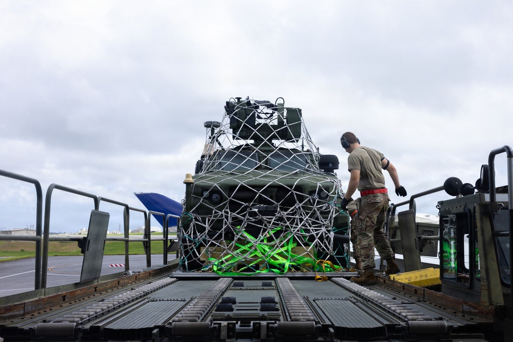A military service member is working on a cargo loading platform, securing or inspecting a military vehicle that is wrapped in a cargo net with bright green and yellow tie-down straps. The scene appears to take place on an airfield tarmac, suggesting the vehicle is being prepared for air transport. The overcast sky and wet-looking surfaces indicate recent or ongoing inclement weather.