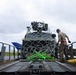 A military service member is working on a cargo loading platform, securing or inspecting a military vehicle that is wrapped in a cargo net with bright green and yellow tie-down straps. The scene appears to take place on an airfield tarmac, suggesting the vehicle is being prepared for air transport. The overcast sky and wet-looking surfaces indicate recent or ongoing inclement weather.
