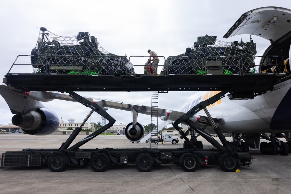 Military cargo loading operation at an airfield, showing personnel using a large scissor-lift loader to transfer netted military equipment onto a commercial or charter cargo aircraft. Two workers in military uniforms are visible on the elevated platform securing or arranging cargo pallets. The aircraft visible has partial markings consistent with a charter or contracted cargo carrier.