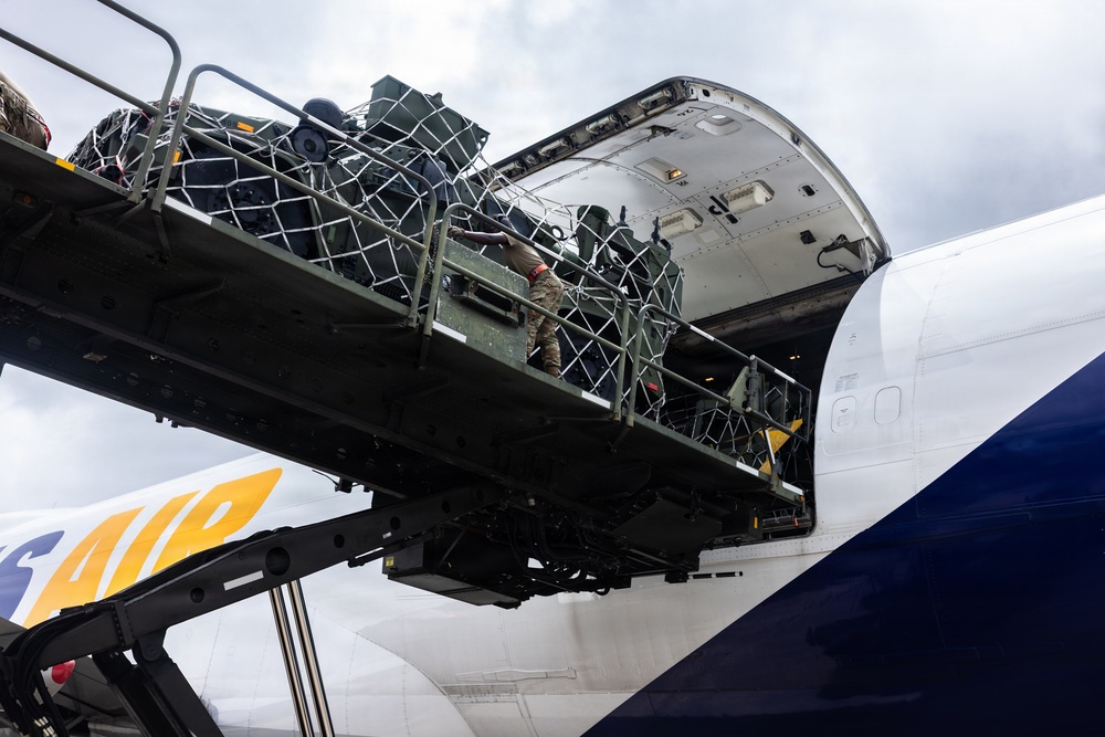 The image shows military cargo being loaded into a large commercial or charter aircraft through its nose or upper deck cargo door. A person in military camouflage uniform is visible on the loading ramp, guiding net-secured cargo into the aircraft. The aircraft livery shows partial text 'AIR' with yellow and blue branding elements.