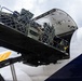 The image shows military cargo being loaded into a large commercial or charter aircraft through its nose or upper deck cargo door. A person in military camouflage uniform is visible on the loading ramp, guiding net-secured cargo into the aircraft. The aircraft livery shows partial text 'AIR' with yellow and blue branding elements.