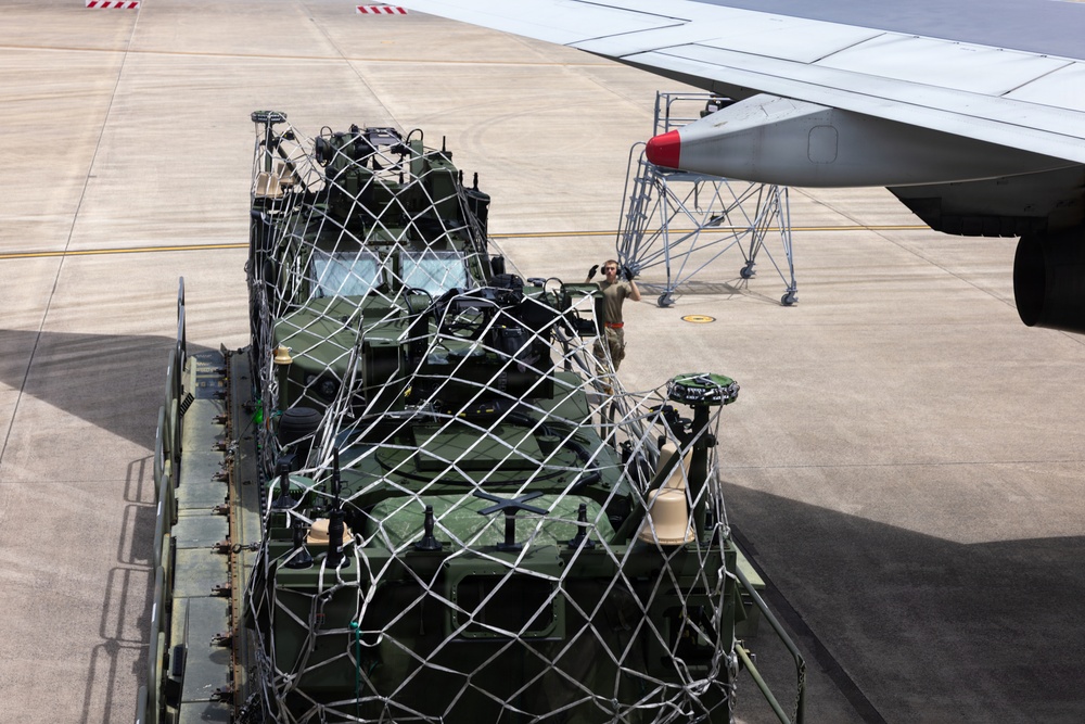 A military wheeled armored vehicle, painted olive green, is secured with cargo netting on a loading platform at an airport tarmac, positioned adjacent to a large aircraft wing. A ground crew member is visible in the background, appearing to signal or communicate. The scene depicts a military airlift or cargo loading operation on a paved airfield.