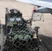 A military wheeled armored vehicle, painted olive green, is secured with cargo netting on a loading platform at an airport tarmac, positioned adjacent to a large aircraft wing. A ground crew member is visible in the background, appearing to signal or communicate. The scene depicts a military airlift or cargo loading operation on a paved airfield.