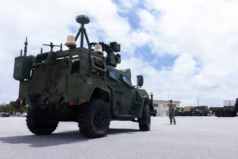 A military tactical vehicle, appearing to be a Joint Light Tactical Vehicle (JLTV) or similar platform, is parked on a paved surface at what appears to be a military installation. The vehicle is equipped with multiple electronic systems, sensors, and communication equipment mounted on its roof. A single service member in camouflage uniform is visible in the background near the vehicle.