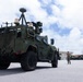 A military tactical vehicle, appearing to be a Joint Light Tactical Vehicle (JLTV) or similar platform, is parked on a paved surface at what appears to be a military installation. The vehicle is equipped with multiple electronic systems, sensors, and communication equipment mounted on its roof. A single service member in camouflage uniform is visible in the background near the vehicle.