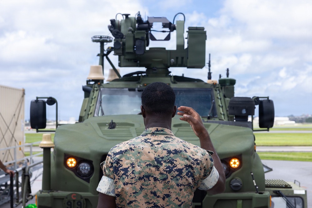 A military service member in MARPAT (Marine pattern) camouflage uniform is seen from behind, gesturing with one hand toward a green armored military vehicle equipped with a remote weapon station or turret system mounted on top. The vehicle appears to be a Joint Light Tactical Vehicle (JLTV) or similar modern armored platform with amber running lights illuminated. The setting appears to be an airfield or military installation tarmac under a partly cloudy sky.