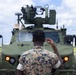 A military service member in MARPAT (Marine pattern) camouflage uniform is seen from behind, gesturing with one hand toward a green armored military vehicle equipped with a remote weapon station or turret system mounted on top. The vehicle appears to be a Joint Light Tactical Vehicle (JLTV) or similar modern armored platform with amber running lights illuminated. The setting appears to be an airfield or military installation tarmac under a partly cloudy sky.