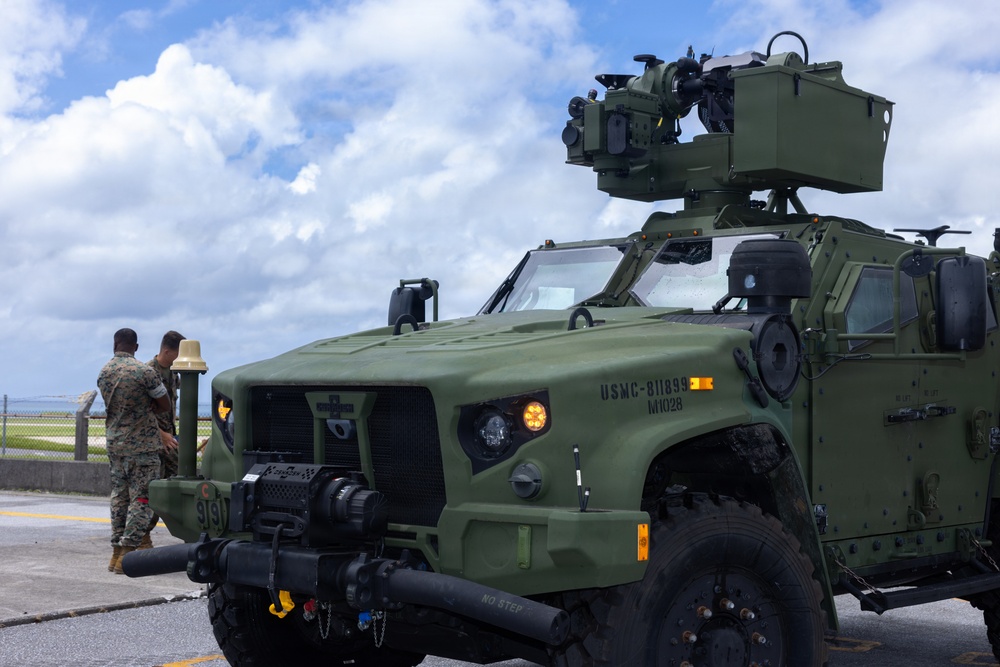 A U.S. Marine Corps Joint Light Tactical Vehicle (JLTV) bearing the markings 'USMC-811899' and 'M1028' is parked on a paved surface, equipped with a roof-mounted weapons system. Two Marines in camouflage uniforms stand near a post in the background. The setting appears to be a coastal military installation based on the visible water and infrastructure in the background.