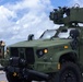 A U.S. Marine Corps Joint Light Tactical Vehicle (JLTV) bearing the markings 'USMC-811899' and 'M1028' is parked on a paved surface, equipped with a roof-mounted weapons system. Two Marines in camouflage uniforms stand near a post in the background. The setting appears to be a coastal military installation based on the visible water and infrastructure in the background.
