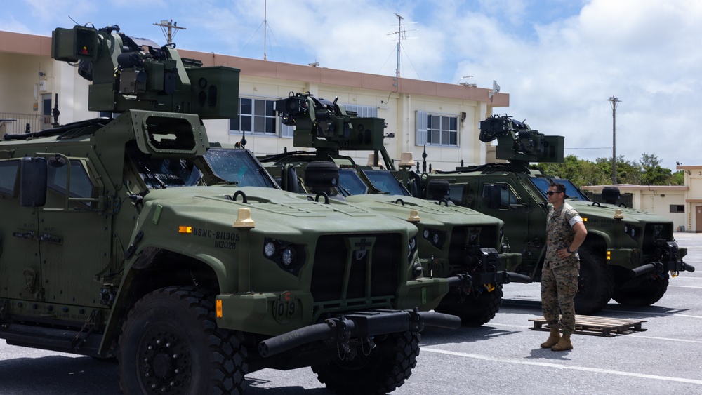 A row of olive-green military tactical vehicles, identifiable as Joint Light Tactical Vehicles (JLTVs), are parked in a paved lot at what appears to be a U.S. Marine Corps installation. The vehicles are equipped with roof-mounted weapon systems and communications equipment. A single service member in camouflage uniform stands beside the vehicles, appearing to inspect or observe them.