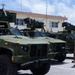 A row of olive-green military tactical vehicles, identifiable as Joint Light Tactical Vehicles (JLTVs), are parked in a paved lot at what appears to be a U.S. Marine Corps installation. The vehicles are equipped with roof-mounted weapon systems and communications equipment. A single service member in camouflage uniform stands beside the vehicles, appearing to inspect or observe them.