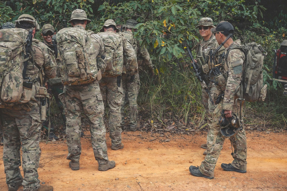 A group of military personnel in camouflage uniforms are gathered on a dirt path near dense jungle vegetation. They are equipped with tactical gear including large rucksacks, weapons, and body armor. The group appears to be in a briefing or rest pause during a field training exercise or patrol operation.