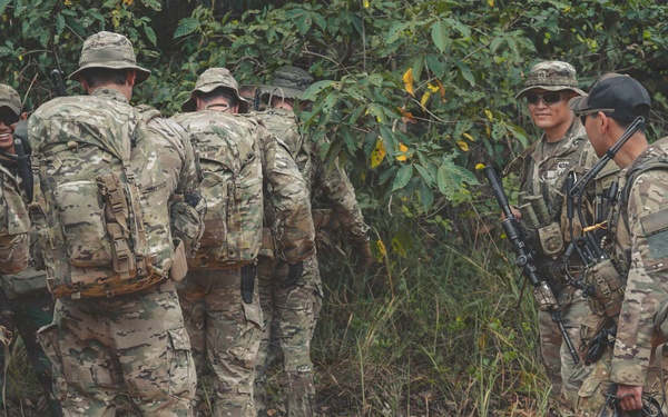 A group of military personnel in camouflage uniforms are gathered on a dirt path near dense jungle vegetation. They are equipped with tactical gear including large rucksacks, weapons, and body armor. The group appears to be in a briefing or rest pause during a field training exercise or patrol operation.