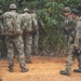 A group of military personnel in camouflage uniforms are gathered on a dirt path near dense jungle vegetation. They are equipped with tactical gear including large rucksacks, weapons, and body armor. The group appears to be in a briefing or rest pause during a field training exercise or patrol operation.