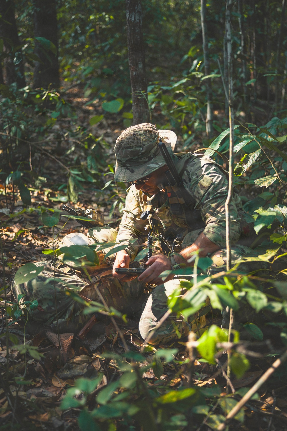 A soldier in full camouflage gear is crouched low in a dense jungle or forest environment. The individual appears to be operating a handheld radio or communication device while positioned among undergrowth and leaf litter. The scene suggests a military field exercise or operational patrol in a tropical woodland setting.