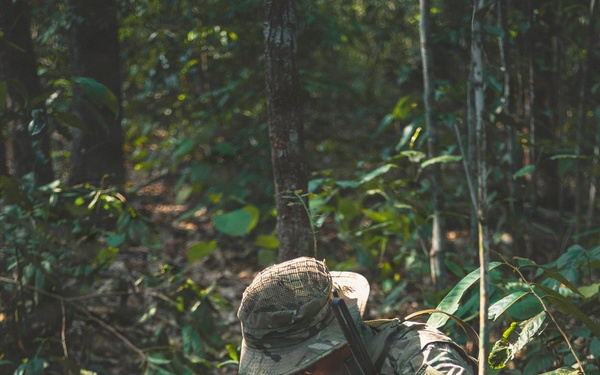 A soldier in full camouflage gear is crouched low in a dense jungle or forest environment. The individual appears to be operating a handheld radio or communication device while positioned among undergrowth and leaf litter. The scene suggests a military field exercise or operational patrol in a tropical woodland setting.