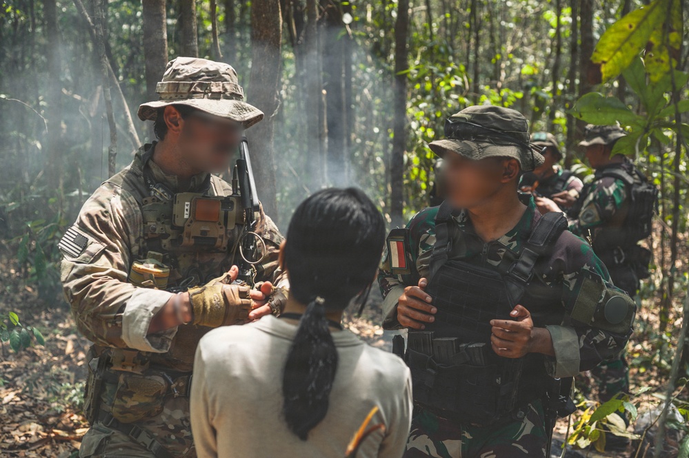 The image shows what appears to be a joint military training exercise in a forested environment. A soldier in US military multicam uniform with an American flag patch is interacting with personnel wearing Indonesian military (TNI) camouflage uniforms. A civilian individual with their back to the camera is present between the two military personnel, suggesting a possible interpreter or role-player scenario.