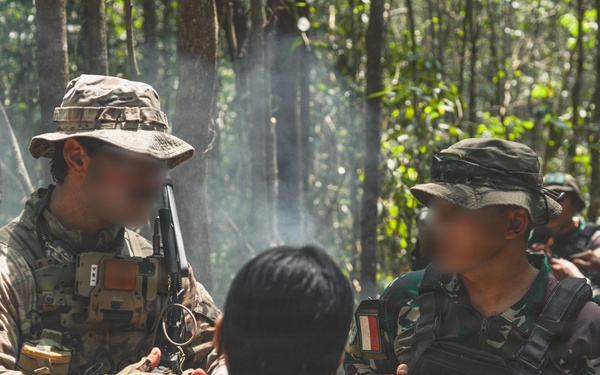 The image shows what appears to be a joint military training exercise in a forested environment. A soldier in US military multicam uniform with an American flag patch is interacting with personnel wearing Indonesian military (TNI) camouflage uniforms. A civilian individual with their back to the camera is present between the two military personnel, suggesting a possible interpreter or role-player scenario.