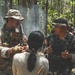 The image shows what appears to be a joint military training exercise in a forested environment. A soldier in US military multicam uniform with an American flag patch is interacting with personnel wearing Indonesian military (TNI) camouflage uniforms. A civilian individual with their back to the camera is present between the two military personnel, suggesting a possible interpreter or role-player scenario.