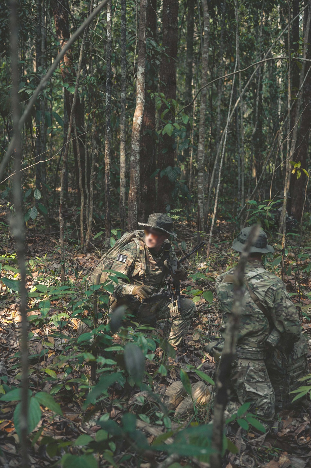 Two armed personnel in full camouflage gear are moving through a dense tropical or subtropical forest environment. Both individuals wear boonie hats and carry rifles, with one displaying what appears to be a US flag patch on their sleeve. The setting and equipment suggest a military patrol, training exercise, or special operations activity in a jungle terrain.