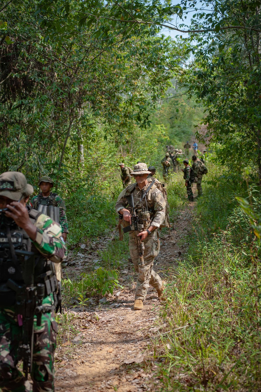 A mixed group of military personnel conducting a patrol along a narrow dirt trail through dense tropical jungle vegetation. The group includes soldiers in at least two distinct camouflage patterns, suggesting a multinational joint training exercise or operation. The central figure wears tan/multicam uniform with a boonie hat and carries a rifle with tactical accessories.